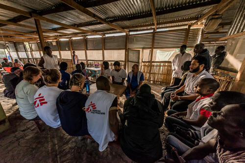 Daily morning meeting of the entire MSF medical team, in a large room of the hospital, a wooden structure covered with a tarpaulin. Everyone is required to participate: doctors, nurses, translators, etc. The program of the day is detailed and questions are asked and answered.

Réunion matinale quotidienne de toute l’équipe médicale de MSF, dans une grande pièce de l'hôpital, une structure en bois recouverte d’une bâche. Tout le monde est tenu d’y participer : médecins, infirmières,  traducteurs, etc… Le programme de la journée y est détaillée et les questions relatives à la marche de la clinique posées.