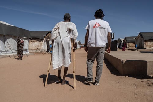 TAWILA, North Darfur, Sudan — March 2026: A man injured during fighting in El Fasher in October 2025 walks on crutches outside the MSF hospital in Tawila, escorted by Hussein, a longtime Médecins Sans Frontières (MSF) medical coordinator. Tawila has become a critical refuge for hundreds of thousands fleeing violence in and around the besieged city of El Fasher, where humanitarian organizations such as MSF provide lifesaving medical care to war-wounded civilians and severely malnourished children. (Photo by Giles Clarke/OCHA)