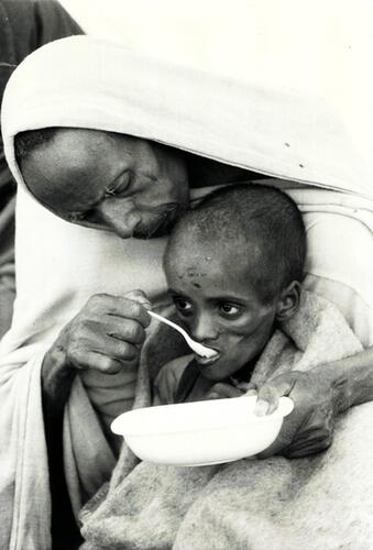 Pere donnant a manger a son enfant malnutri. Le conflit avec la Somalie, la guerre de secession erythreenne, le soulevement Tigrayen, ont epuise la plus grande partie des ressources de ce pays. Chasses par la famine, des centaines de milliers de personnes ont quitte leurs villages d’Erythree, du Tigray, du Wollo, pour gagner les centres de distribution de nourriture ou les camps de refugies du Soudan. MSF intervient dans les camps de Korem et de Kobo, dans la province du Wollo: des centres de recuperation nutritionnelle ont ete mis en place, complement indispensable des salles d’hospitalisation et de convalescence, de la maternite, du laboratoire et des centres de consultation.